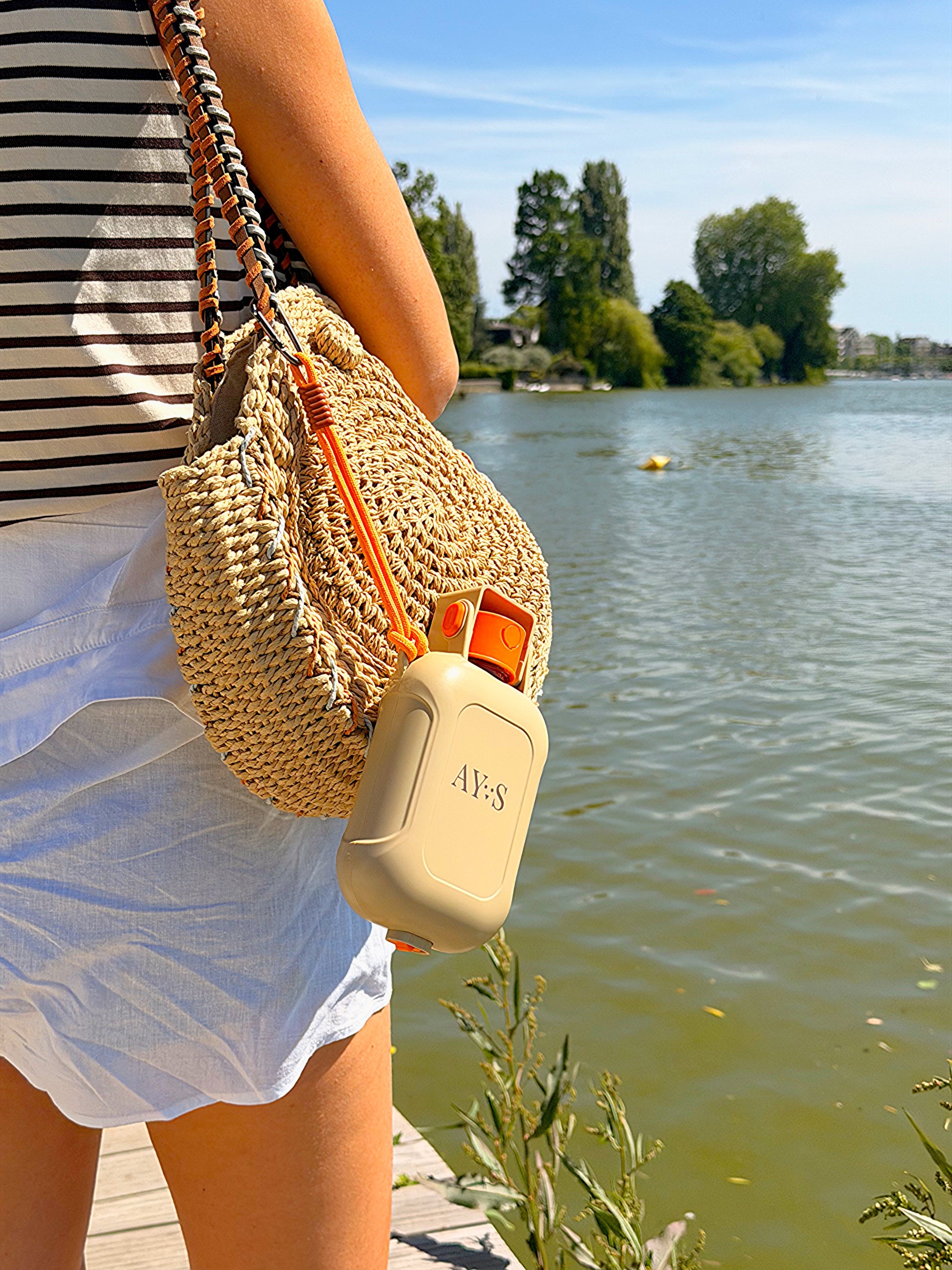 Gourde de voyage pour chien AYOS 2-en-1 suspendue à un sac en osier au bord d’un lac. Accessoire portable avec réservoir d’eau et gamelle intégrée, parfait pour les promenades, randonnées, vacances et activités en extérieur avec son chien.