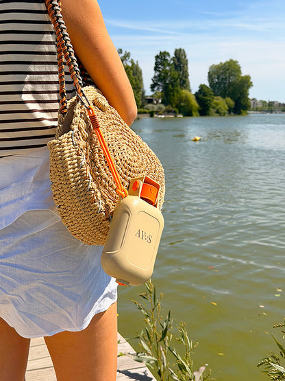 Gourde de voyage pour chien AYOS 2-en-1 suspendue à un sac en osier au bord d’un lac. Accessoire portable avec réservoir d’eau et gamelle intégrée, parfait pour les promenades, randonnées, vacances et activités en extérieur avec son chien.