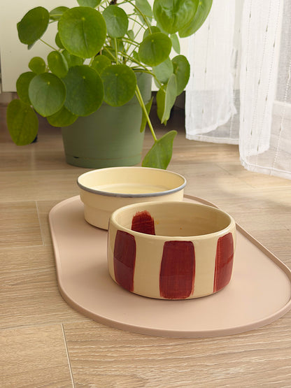 Tapis de gamelle AYOS en silicone couleur grège avec deux gamelles posé sur un sol clair près d’une plante verte et d’une fenêtre.