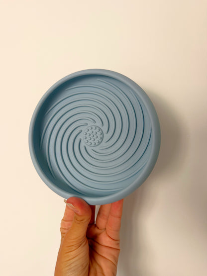 Le Nuage Bowl bleu ciel, tapis de léchage en silicone pour chien, tenu en main afin d’illustrer sa taille et sa texture spiralée favorisant un léchage lent et apaisant.